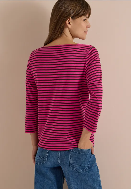 Frau von hinten in Jeans, trägt pink-schwarz gestreiftes 3/4-Arm-Shirt und schaut seitlich, vor hellem Hintergrund.