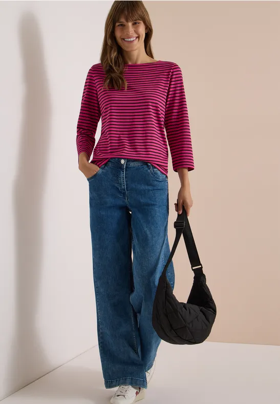 Frau in pink-gestreiftem Oberteil und weiter Jeans, trägt schwarze Tasche vor hellem Hintergrund.