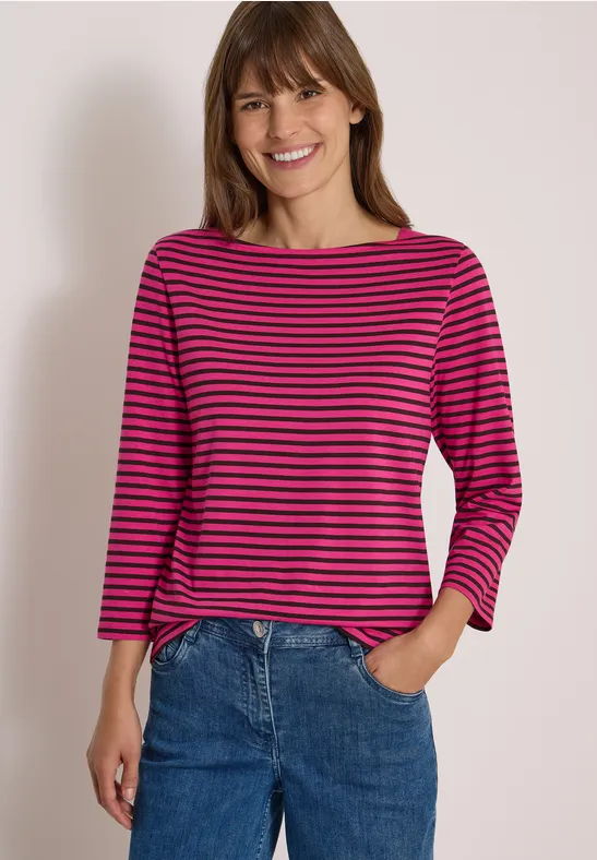 Eine Frau mit pink-blau gestreiftem Shirt und Jeans lächelt in die Kamera.