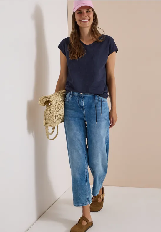 Frau in lässigem Outfit: dunkelblaues T-Shirt, weite Jeans, rosa Mütze, Strohtasche, braune Schuhe
