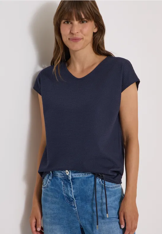 Frau trägt dunkelblaues T-Shirt mit kurzen Ärmeln und Jeans, lächelt leicht.