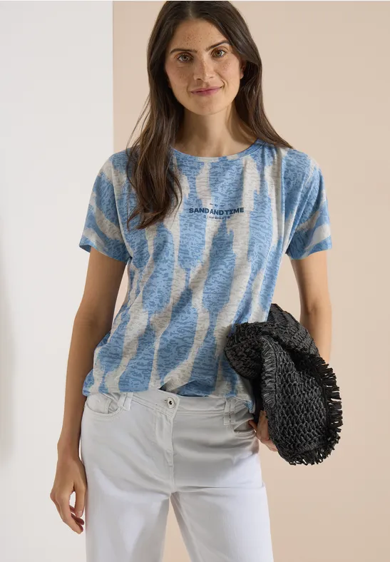 Femme en t-shirt bleu-blanc et pantalon blanc tenant un sac noir devant un mur beige.