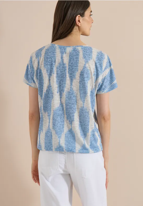 Femme de dos portant un pull court à motifs bleus et blancs et un pantalon blanc sur fond clair.
