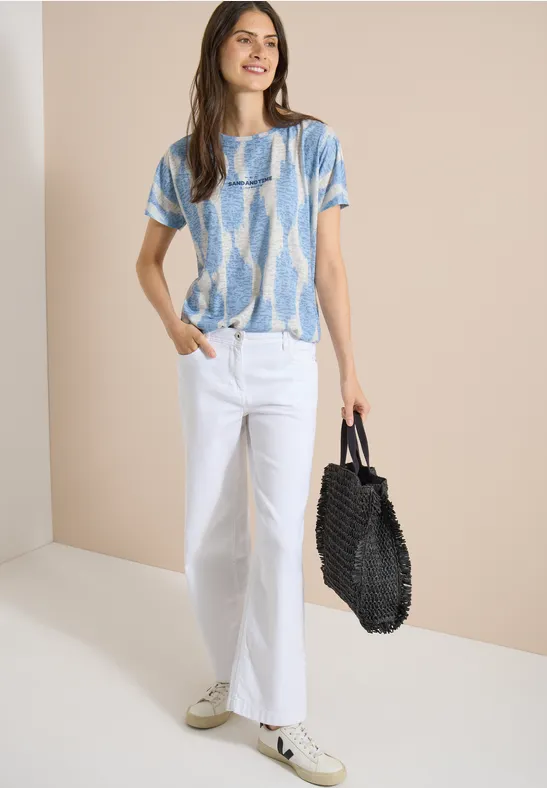 Femme portant un t-shirt bleu et blanc, un jean blanc, des baskets et un sac noir sur fond neutre.