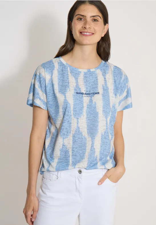 Femme portant un t-shirt bleu et blanc à motifs et un jean blanc, devant un fond clair, la main dans la poche.