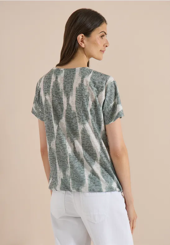 Femme de dos aux cheveux lâchés portant un top ample à motif vert et blanc et un pantalon blanc.