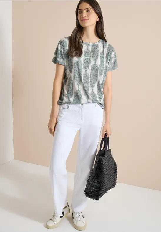 Femme en jean blanc, blouse imprimé serpent et baskets, portant un grand sac noir tressé sur fond beige.