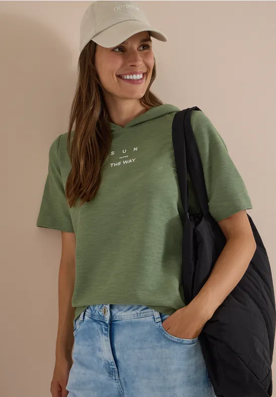 Eine lächelnde Frau in grüner Bluse, Jeans und Cap mit schwarzer gesteppter Schultertasche.