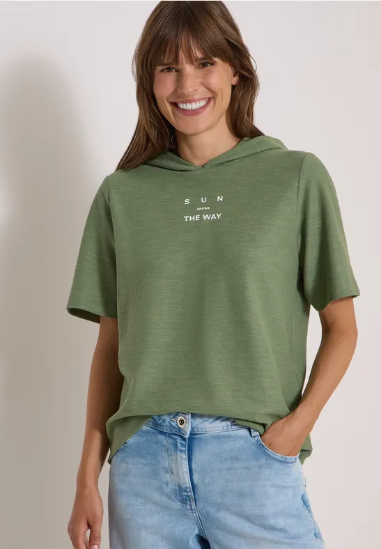 Frau mit kurzem, grünem Kapuzen-Shirt und hellblauen Jeans, lächelnd mit Händen in den Taschen.