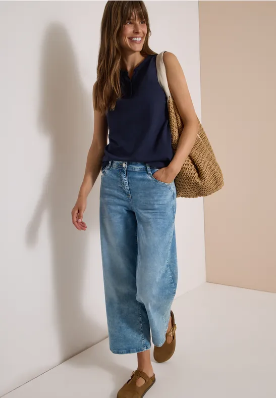 Vrouw leunt tegen lichte muur, draagt donkerblauw topje, wijde jeans, stro-tas en bruine schoenen.