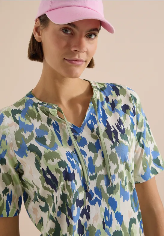 Person trägt kurzärmliges Kleid mit grün-blau-weißem abstraktem Muster und eine rosa Kappe.