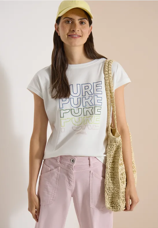 Frau mit weißem T-Shirt 'PURE', rosa Hose und gehäkelter Strandtasche.