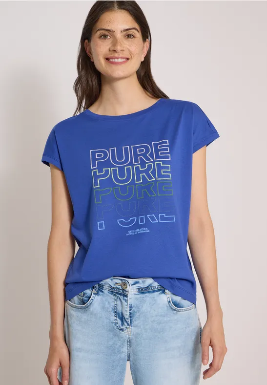 Frau in blauem T-Shirt mit Aufdruck "PURE PURE PURE" und hellen Jeans vor schlichtem Hintergrund.