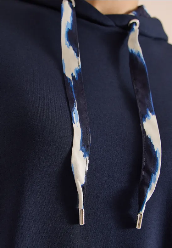 Nahaufnahme eines dunkelblauen Hoodies mit weiß-blau gemusterten Kordeln und silbernen Metallenden.