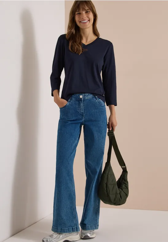 Frau in dunkelblauem Shirt, Schlagjeans und olivgrünem Beutel vor hellem Hintergrund.