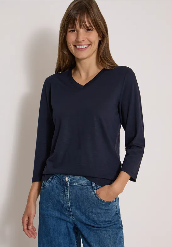 Fröhliche Frau trägt dunkelblauen V-Pullover mit 3/4-Ärmeln und blaue Jeans.