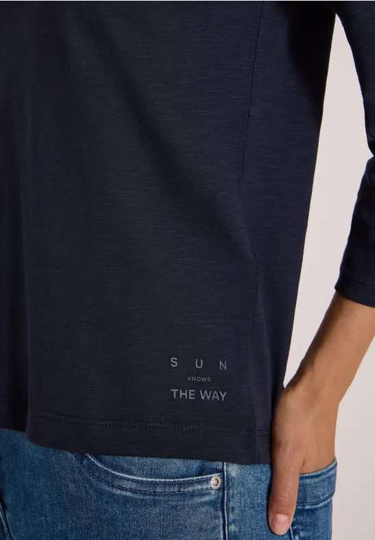 Eine Person trägt ein dunkelblaues 3/4-ärmeliges Shirt mit Schriftzug "SUN THE WAY" und steckt die Hand in die Jeans.