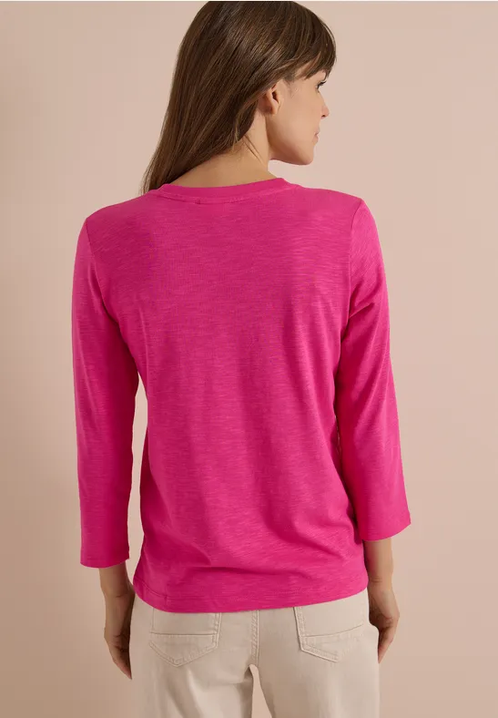 Eine Frau von hinten in einem pinken 3/4-Arm-Shirt und beigen Hosen.