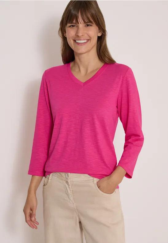 Frau im pinken Langarmshirt mit V-Ausschnitt und beigen Hosen, lächelnd, Hand in Tasche.