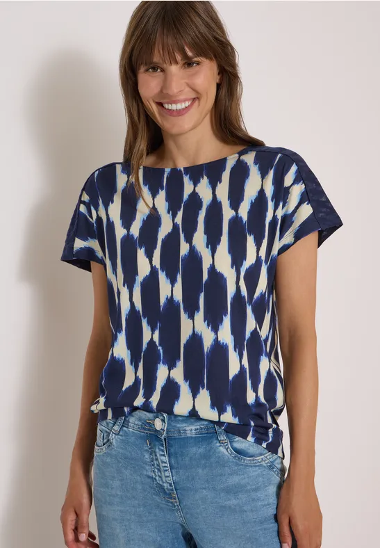 Fröhliche Frau im blau-weiß gemusterten Kurzarmshirt und Jeans vor hellem Hintergrund.