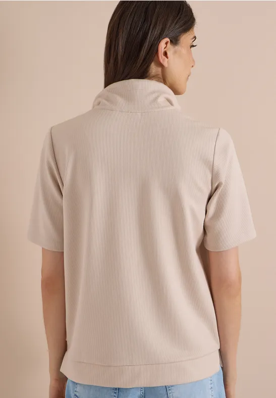 Rückansicht einer Frau in einem beigen Kurzarmshirt mit Stehkragen vor neutralem Hintergrund.