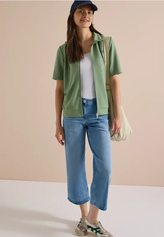 Vrouw in wit shirt, groene blouse, blauwe cropped jeans, espadrilles en schoudertas tegen neutrale achtergrond.