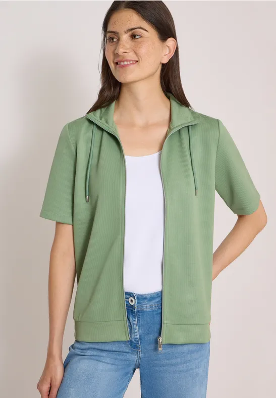 Model draagt groene korte mouw hoodie met rits, wit topje en blauwe jeans.