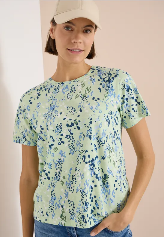Eine Person trägt ein kurzärmeliges T-Shirt mit grün-blauem Muster und steht mit einer Hand in der Hosentasche.