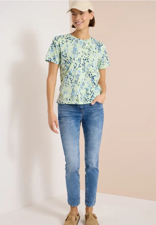 Frau im Stehen trägt grün geblümtes T-Shirt, blaue Jeans und weiße Sneaker vor hellem Hintergrund.