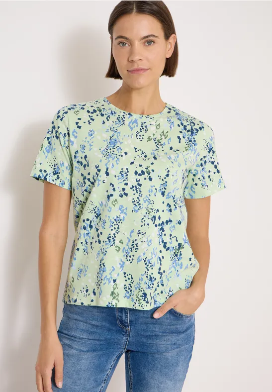 Dame trägt grünes T-Shirt mit blauem Blumenmuster und Jeans vor hellem Hintergrund.