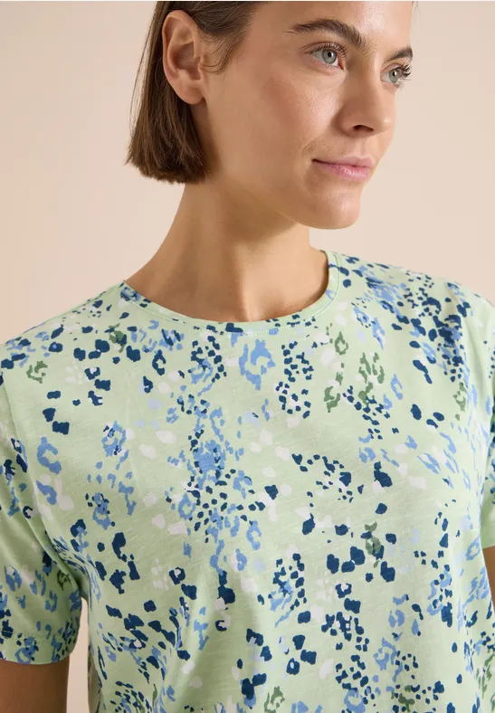 Eine Frau in einem hellgrünen T-Shirt mit abstraktem blauem Fleckenmuster, schaut seitlich.