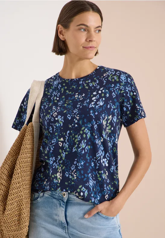 Frau trägt blaues T-Shirt mit Blumenmuster, halb in Jeans gesteckt, mit Strohtasche über der Schulter.