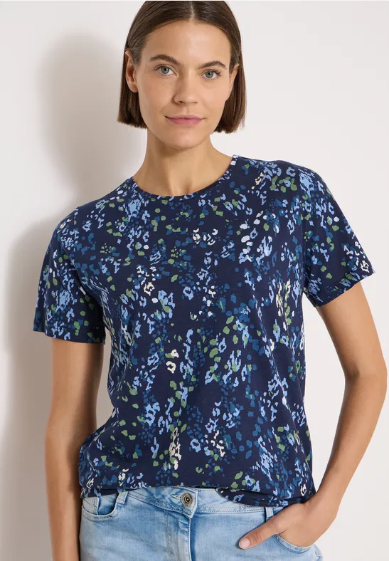 Frau trägt dunkelblaues, kurzärmeliges Shirt mit Blumenmuster und helle Jeans vor weißem Hintergrund