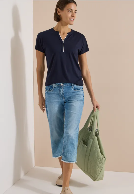 Frau in blauem T-Shirt mit Reißverschluss, weiten 3/4-Jeans, Beige-Slippern und grünem Steppbeutel.