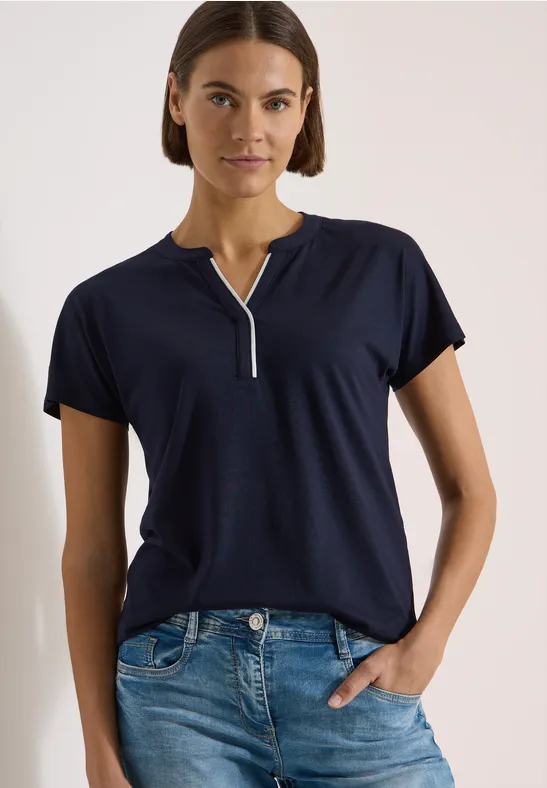 Frau in dunkelblauem Kurzarmshirt mit Knopfleiste und Jeans vor neutralem Hintergrund.