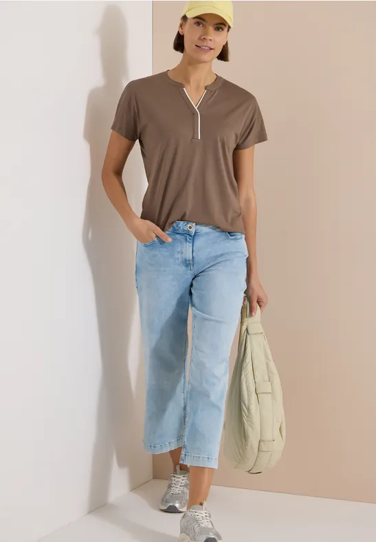 Frau in braunem V-Ausschnitt-Shirt, hellen Jeans und Sneakern, hält beige Tasche, steht vor heller Wand.