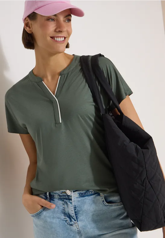 Frau in grünem Kurzarmshirt mit Reißverschluss, Jeans, rosa Cap und großer schwarzer Stepp-Tasche.