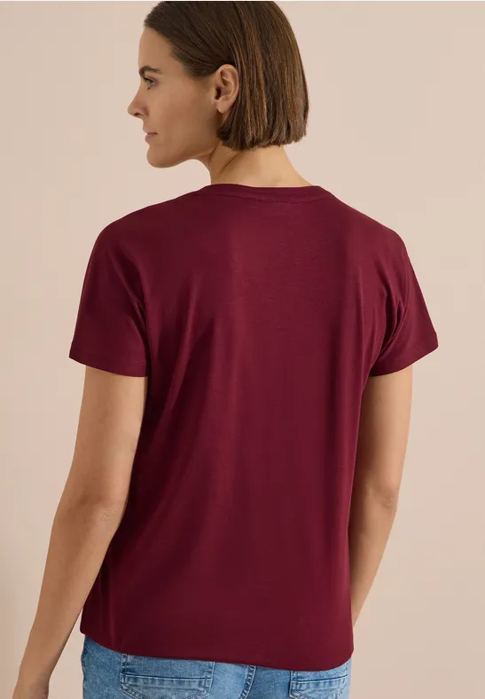 Frau von hinten in schlichtem bordeauxfarbenem T-Shirt mit kurzen Ärmeln, Jeans, neutralem Hintergrund.