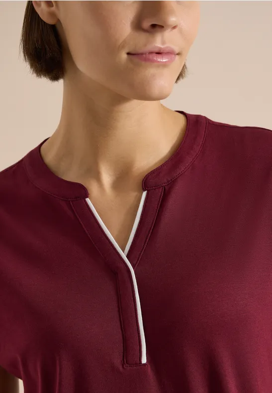 Nahaufnahme einer Person in einem burgunderroten Shirt mit V-förmigem Ausschnitt und weißer Ziernaht.