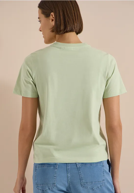 Person steht mit dem Rücken zur Kamera, trägt hellgrünes T-Shirt und blaue Jeans.