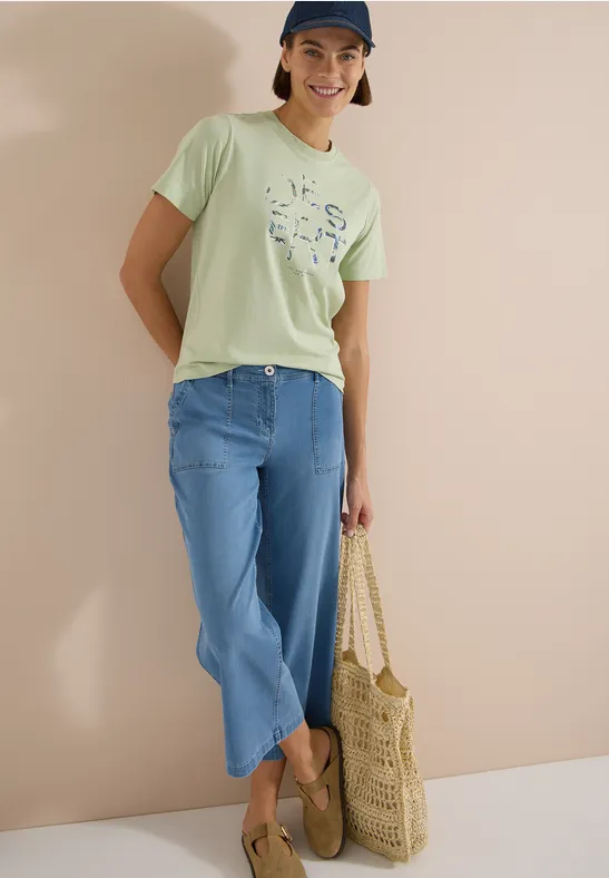 Person trägt hellgrünes Shirt, blaue Jeans, Basttasche und Slipper vor heller Wand.