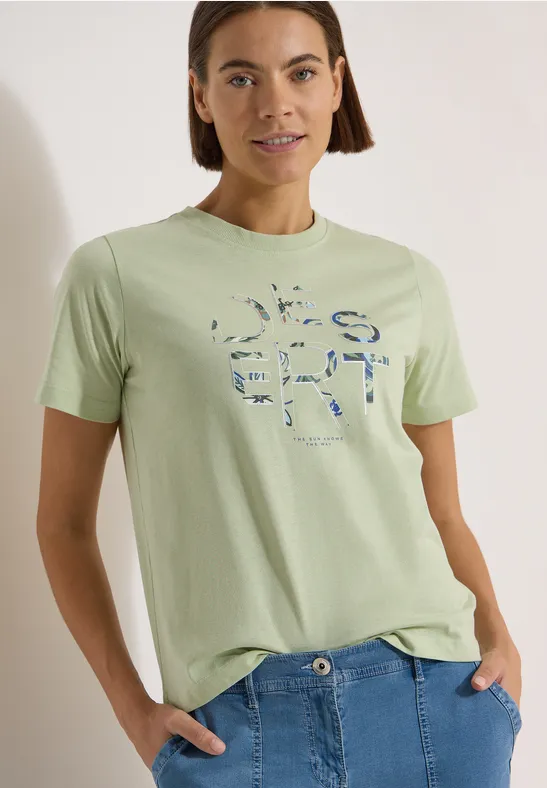 Frau in pastellgrünem T-Shirt mit abstraktem Print und blauen Jeans.