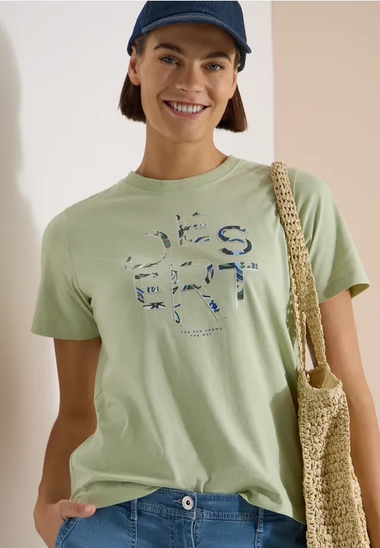 Eine Frau trägt ein hellgrünes T-Shirt mit Print, eine Netz-Tasche über der Schulter und eine dunkle Cap.