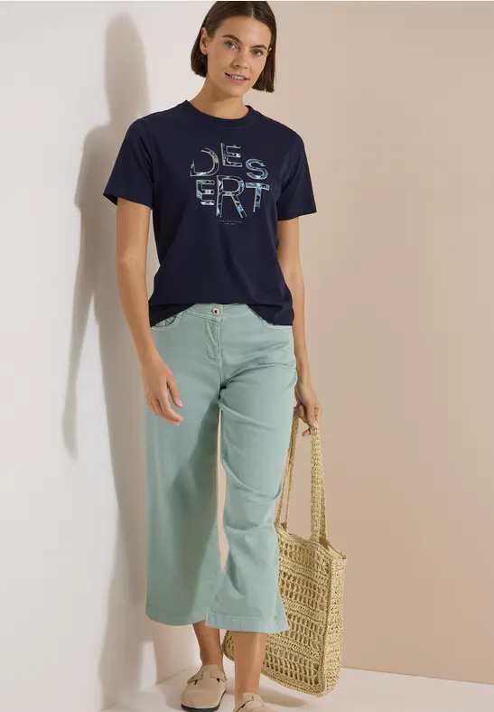 Frau steht vor neutralem Hintergrund, trägt dunkelblaues T-Shirt mit Schriftzug, mintfarbene Culotte und Basttasche.