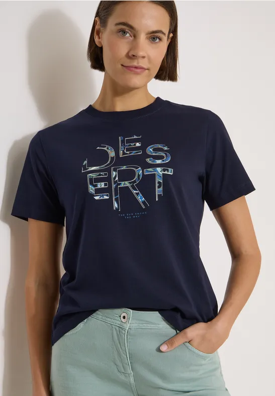 Person trägt dunkelblaues T-Shirt mit ‚DESERT‘-Print und hellgrüner Hose.