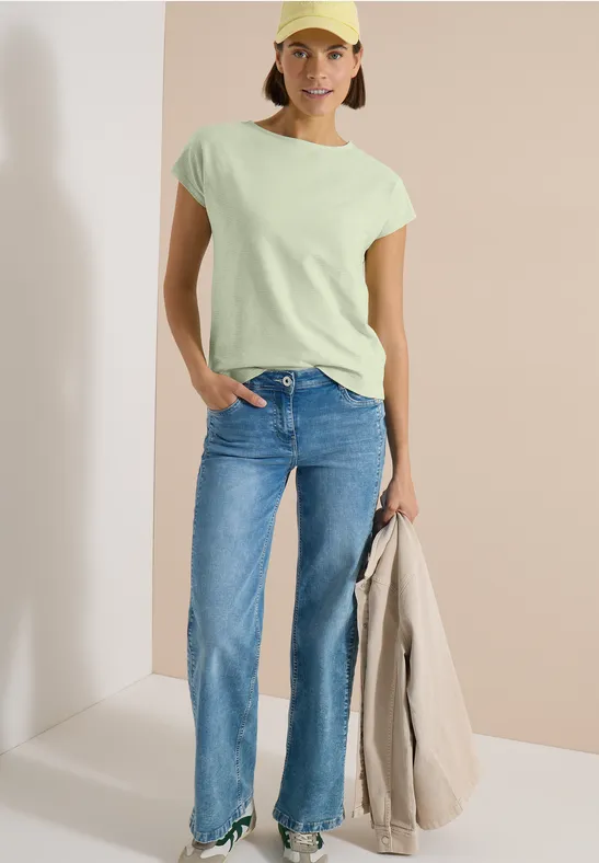 Frau in mintgrünem T-Shirt, blauen geraden Jeans, hält einen beigen Mantel, vor schlichtem Hintergrund.