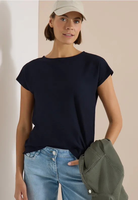 Vrouw draagt donkerblauw T-shirt en spijkerbroek, houdt een jas in haar hand.