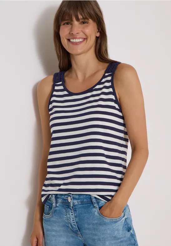 Frau in blau-weiß gestreiftem Tanktop und Jeans, lächelnd mit Händen in den Taschen.