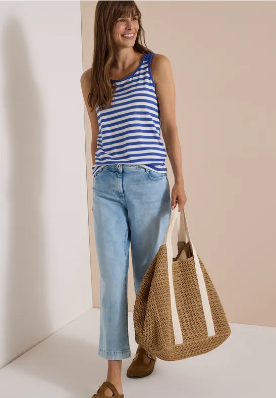 Frau mit gestreiftem Top, blauen Jeans und großem Strohsack, lächelnd vor beigem Hintergrund.