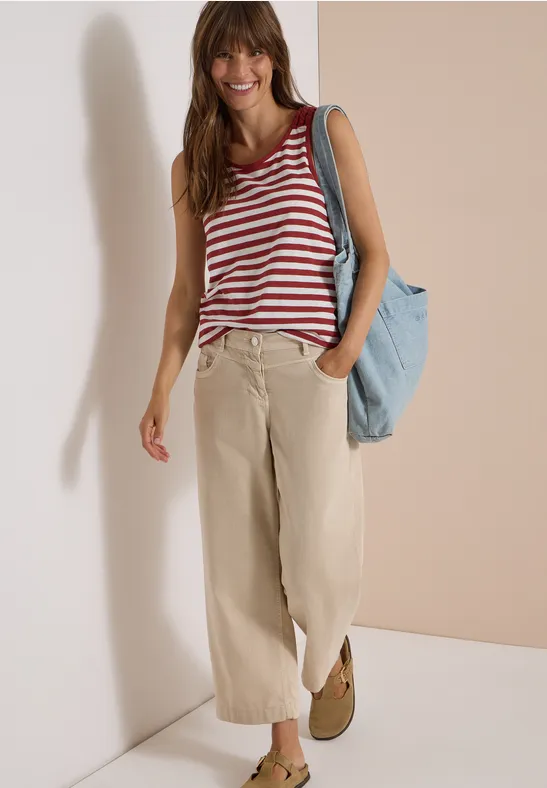 Eine lächelnde Frau in rot-weiß gestreiftem Top, weiten beigen Hosen und mit blauer Tasche vor hellem Hintergrund.